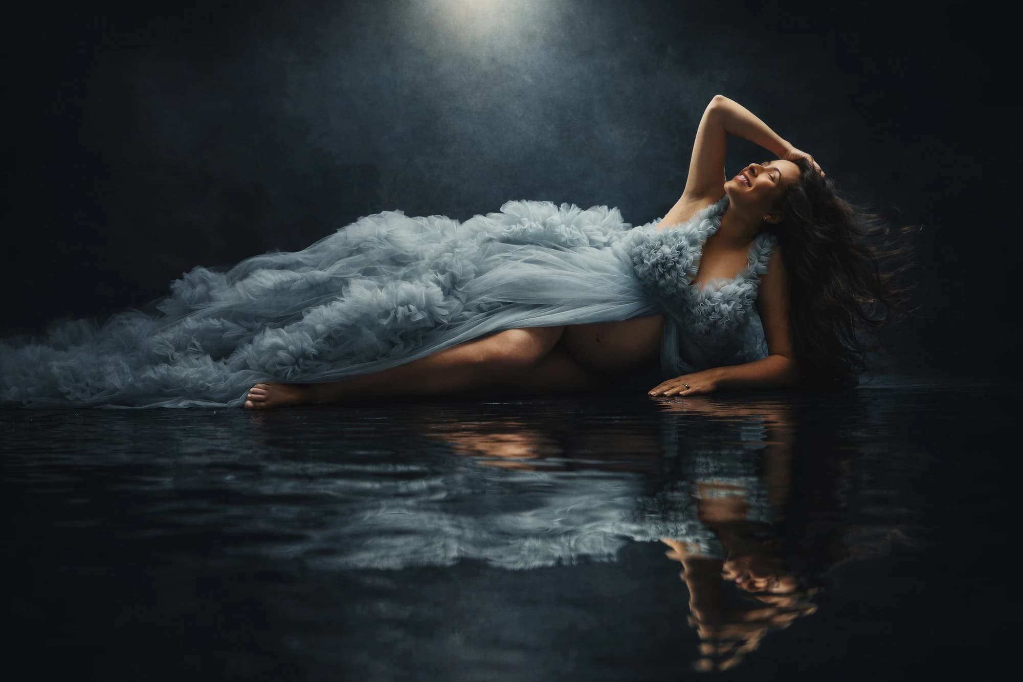 An expectant mother reclining in pale blue ruffles above a dark reflection