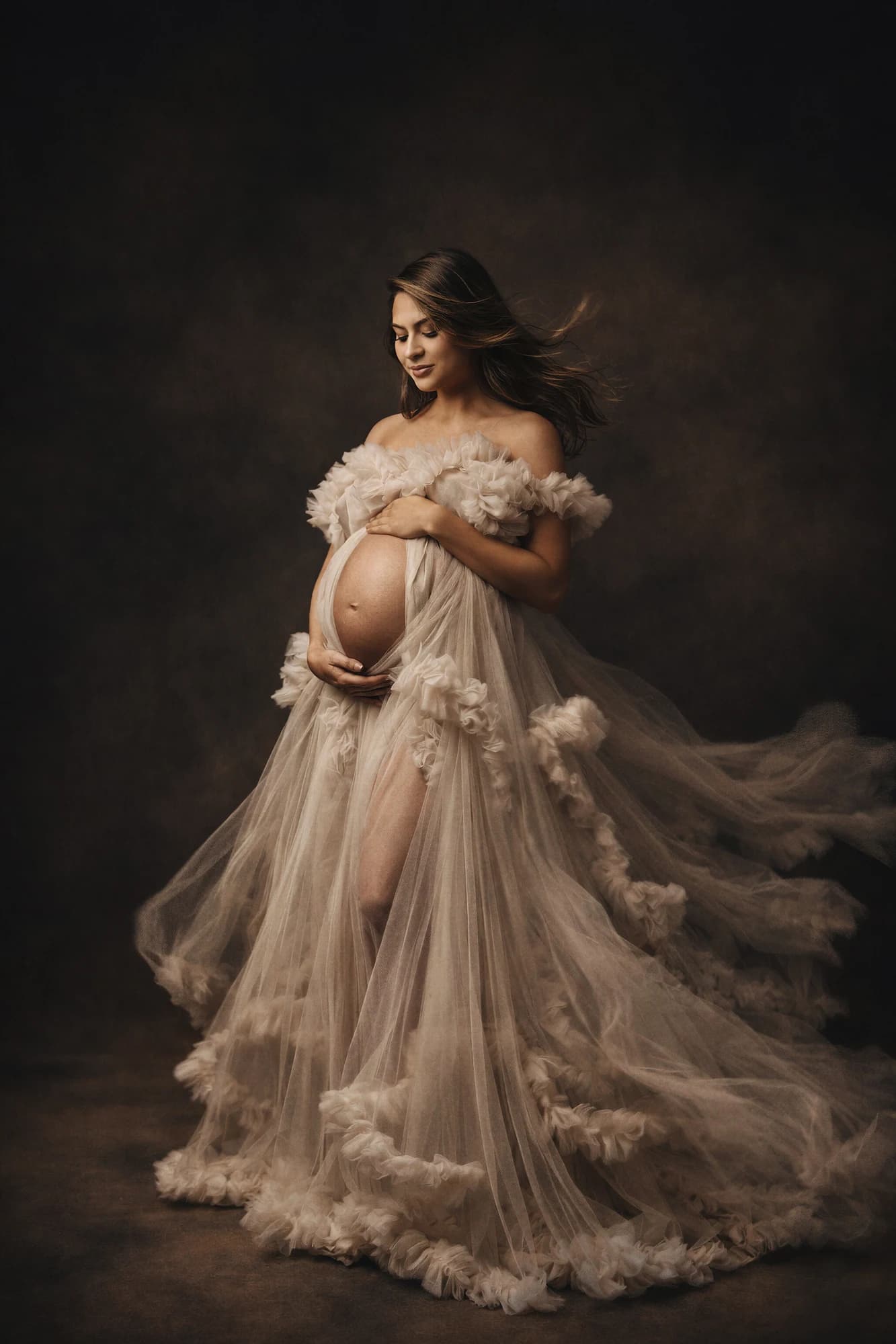 An expectant mother in a flowing champagne tulle gown against a dark backdrop