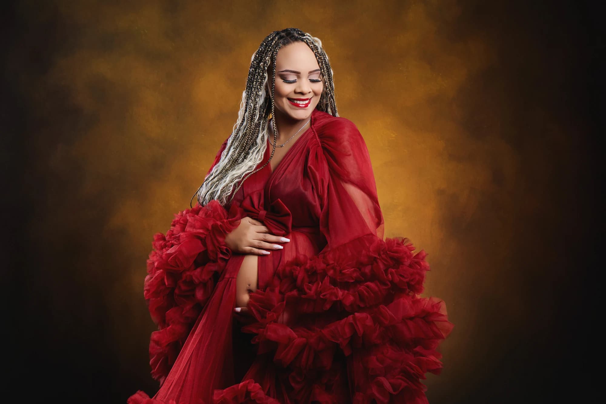 An expectant mother smiling in a red ruffled gown against a golden backdrop