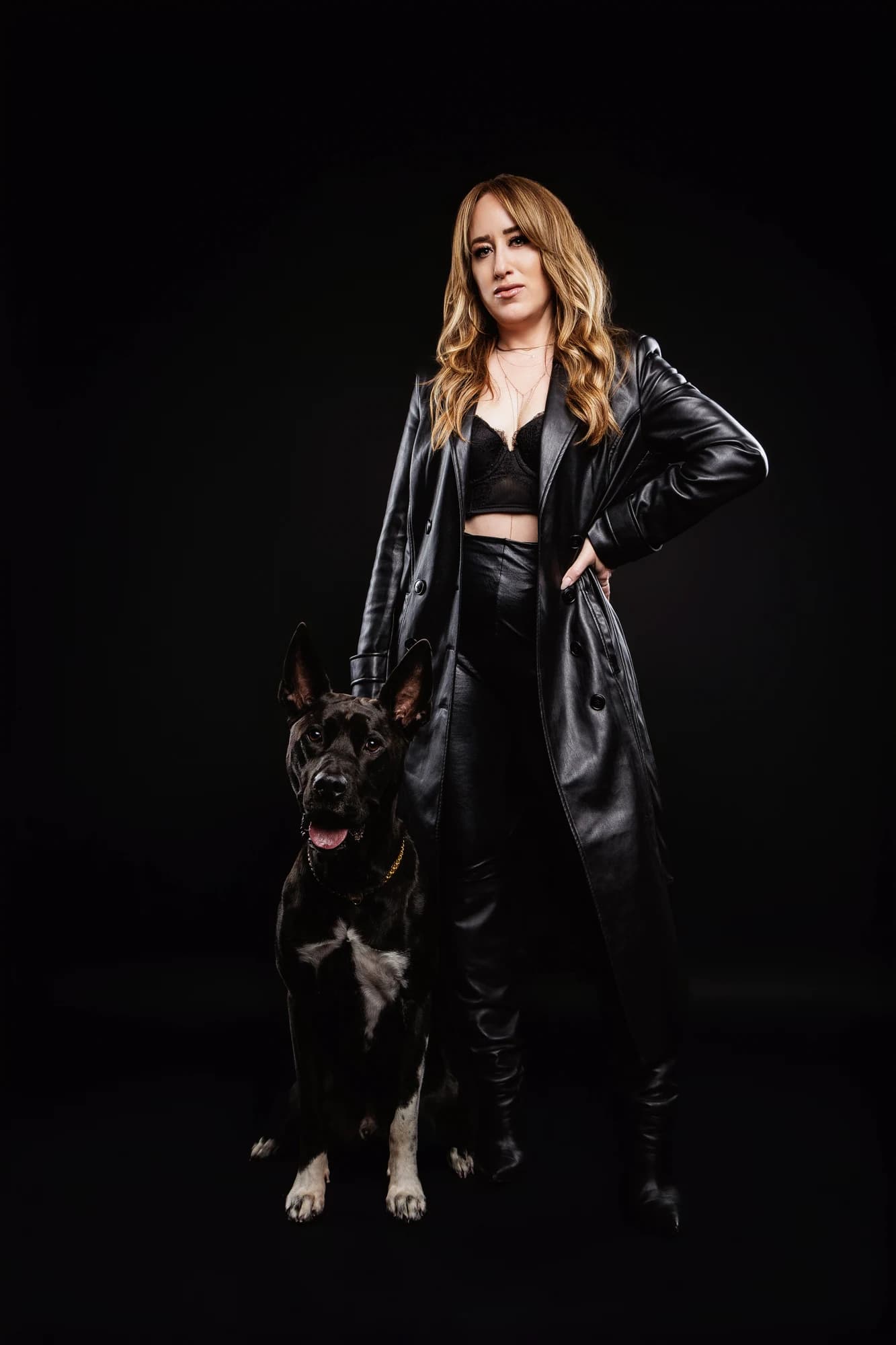 A woman in a long black leather coat standing beside her dog against a dark backdrop