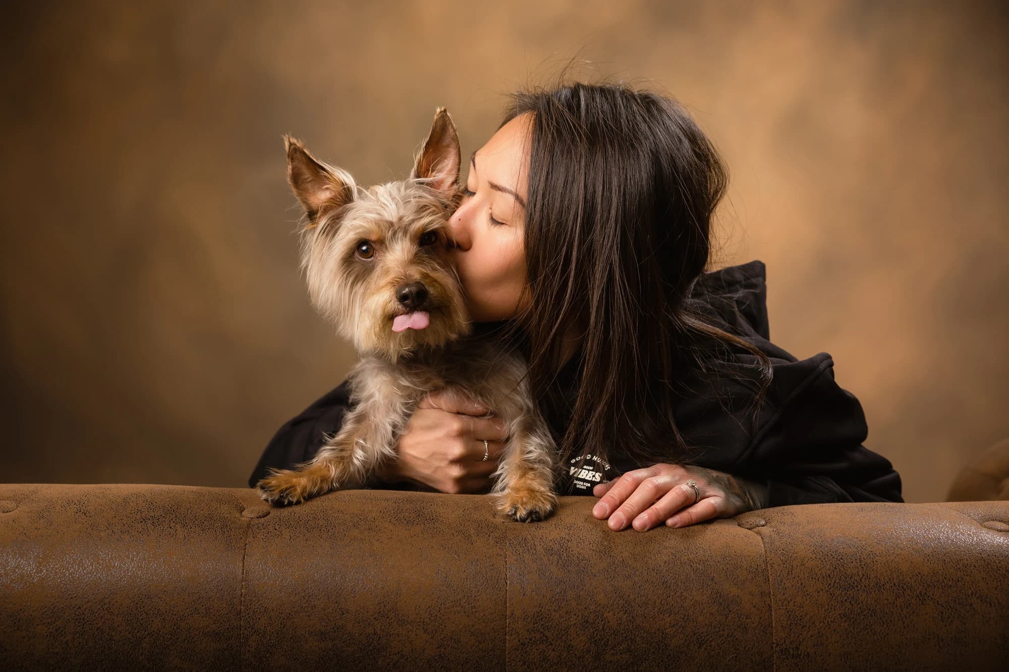 A woman kissing her small terrier as they lean over a leather sofa