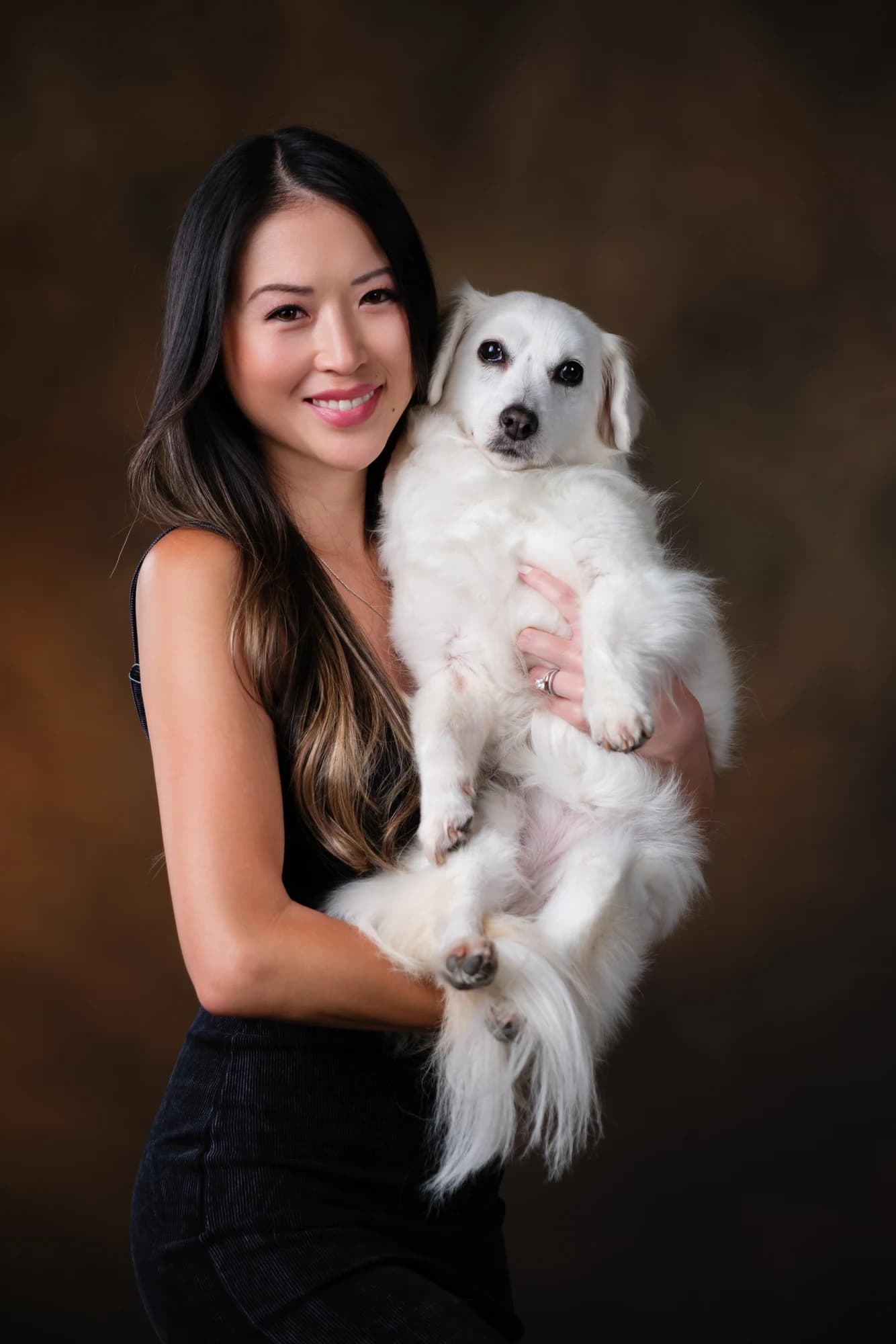 A smiling woman holding her small white dog against a warm studio backdrop