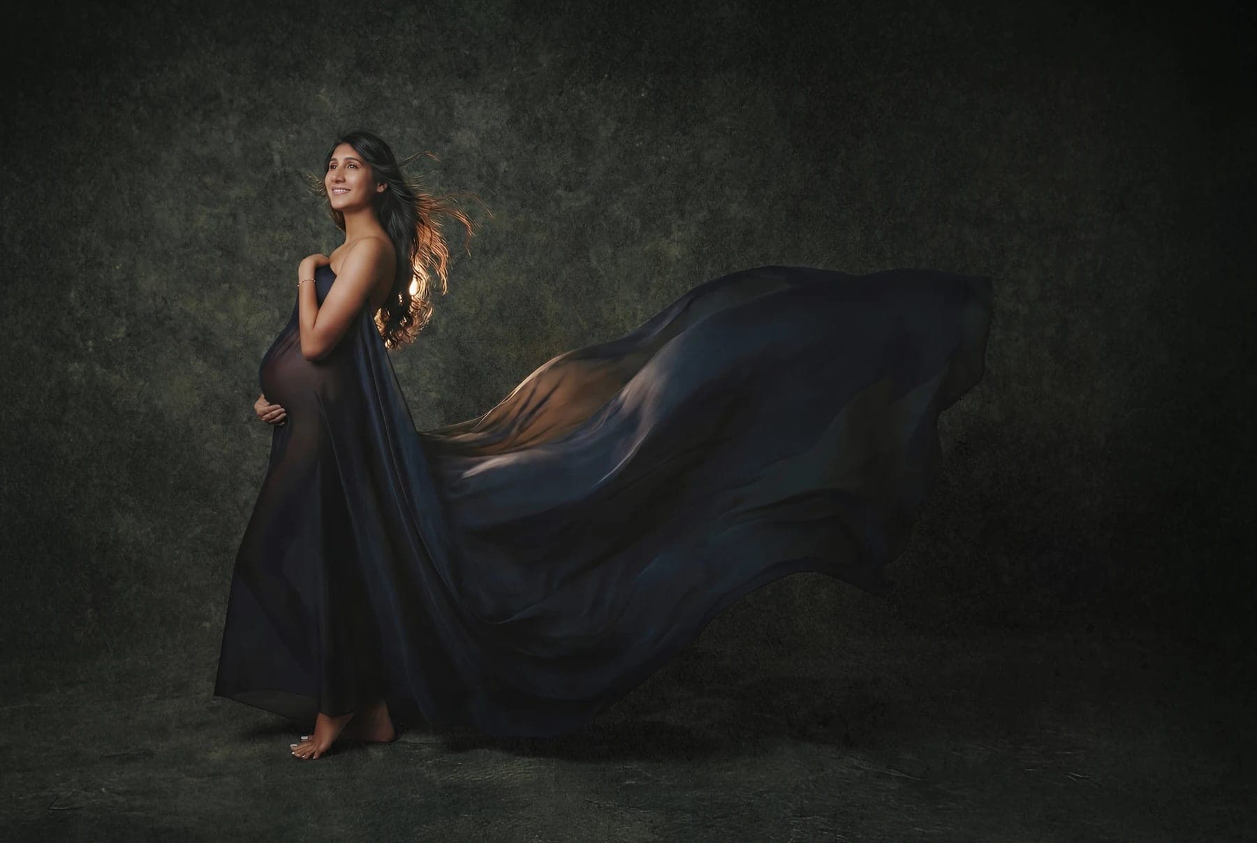 Pregnant woman in flowing dark fabric holds her belly against a mottled green backdrop.