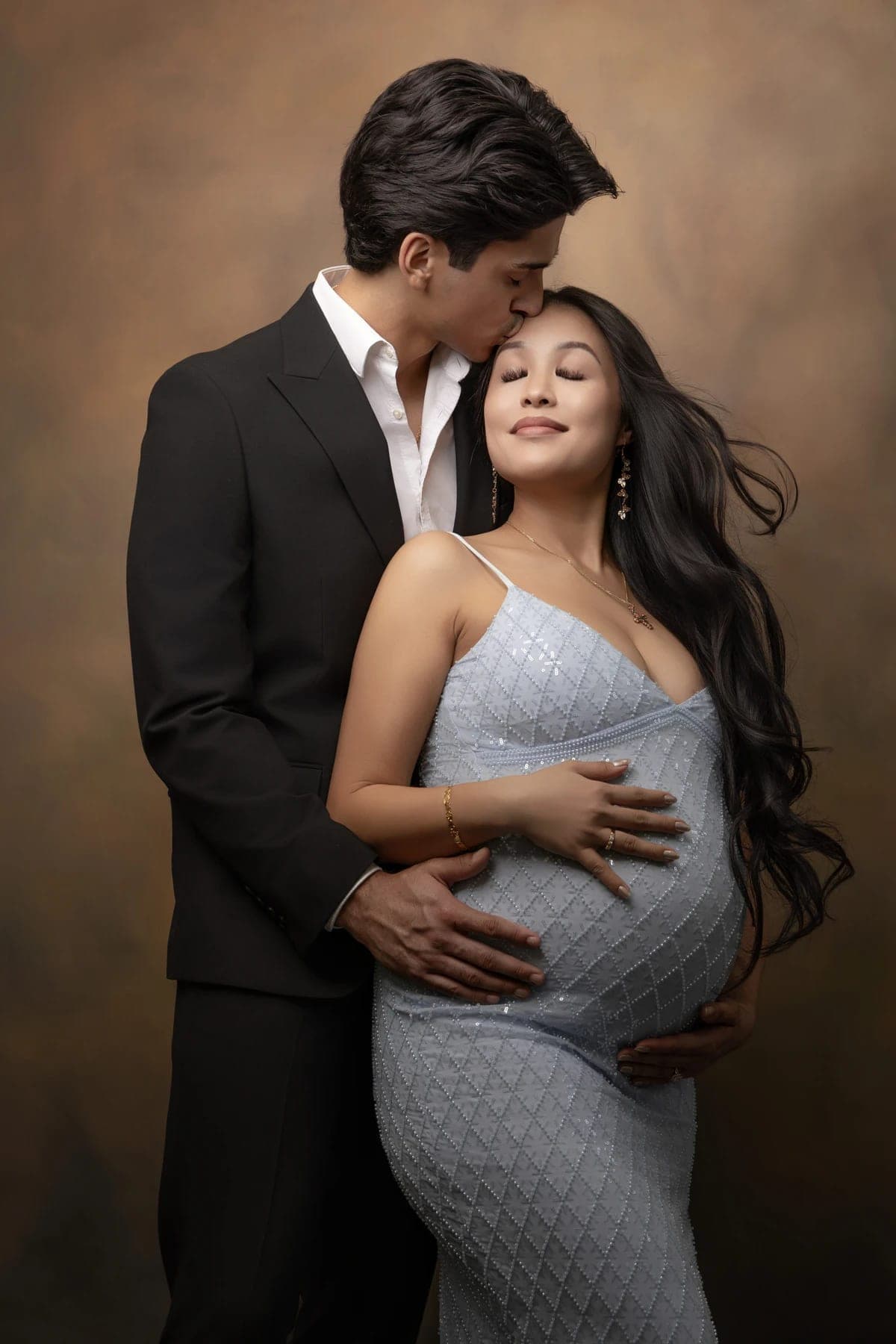 An expectant couple embraces in a blue gown and dark suit against a warm backdrop.